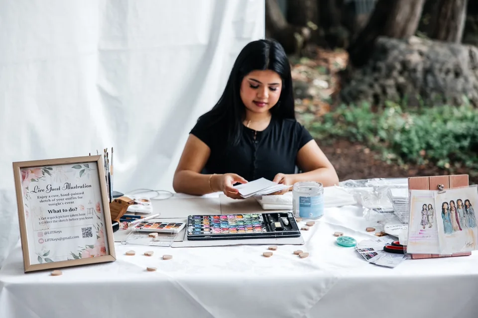 Talia seated at her live illustration table with paints and finished artwork